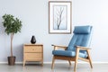 Wooden recliner chair with blue leather cushion near cabinet and side table against white wall with poster frame