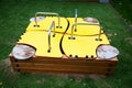 Wooden sandbox covered with bright yellow plywood sheets with stainless steel handles on a background of greenery. Playgrounds