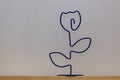 On a wooden shelf, against a background of white wallpaper, stands a figure of a rose flower made of steel wire.