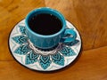 On a wooden table, a blue china cup filled with black coffee, on top of a saucer decorated with a blue and white mandala Royalty Free Stock Photo