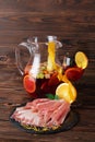 A Plate Of Prosciutto And Ham, A Glass Jug Of Beverage With Apples And Oranges On A Wooden Brown Background.