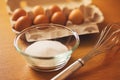 On a wooden table are the ingredients: chicken eggs in a package and sugar in a glass bowl, and next to it is a whisk. Cooking