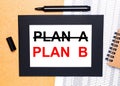 On a wooden table, there is a black open marker, a brown notepad and a sheet of paper in a black frame with the text PLAN B. View