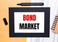 On a wooden table, there is a black open marker, a brown notepad and a sheet of paper in a black frame with the text BOND MARKET.