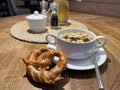 wooden table with a white bowl of broth with cubes of vegetables in and a traditional bavarian brezel on the side