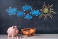 A wooden toy biplane airplane and a piggy bank stand side by side, a chalk board in the background with a picture of the sun and Royalty Free Stock Photo