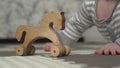 Wooden toy horse stands on the floor. In the background, a small child in a gray and white striped pajamas is playing.