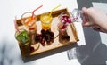 A wooden tray with colorful drinks: orange, yellow, and cucumber lemonade. Nearby are cherries. A hand pours red juice from a