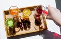 A wooden tray with colorful drinks: orange, yellow, and cucumber lemonade. Nearby are cherries. A hand pours red juice from a