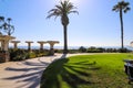 A wooden white pergola surrounded by lush green palm trees and plants with red flowers and lush green grass near blue ocean water Royalty Free Stock Photo