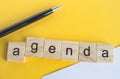 The word agenda is written on wooden cubes between a pen and a white sheet of paper on a yellow background  top view  flat layout Royalty Free Stock Photo