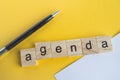 The word agenda is written on wooden cubes between a pen and a white sheet of paper on a yellow background  top view  flat layout Royalty Free Stock Photo