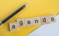 The word agenda is written on wooden cubes between a pen and a white sheet of paper on a yellow background  top view  flat layout Royalty Free Stock Photo