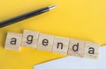 The word agenda is written on wooden cubes between a pen and a white sheet of paper on a yellow background  top view  flat layout Royalty Free Stock Photo