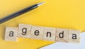 The word agenda is written on wooden cubes between a pen and a white sheet of paper on a yellow background  top view  flat layout Royalty Free Stock Photo