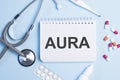 The word aura written on a white notepad on a blue background near a stethoscope, syringe, electronic thermometer and pills.