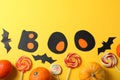 Word Boo, candy, decorative bats and pumpkins on yellow background