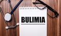 The word BULIMIA is written on white paper on a wooden background near a stethoscope and black-framed glasses. Medical concept Royalty Free Stock Photo