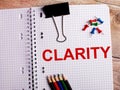 The word CLARITY is written in a notebook near multi-colored pencils and buttons on a wooden background