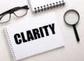 The word CLARITY is written in a white notebook on a white background near a black magnifier and black-framed glasses. Business Royalty Free Stock Photo