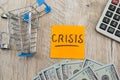 The word `crisis` on a sticker with a shopping cart and dollars on a wooden background. View from above.