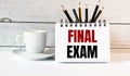 The word FINAL EXAM is written in a white notepad near a white cup of coffee on a light background Royalty Free Stock Photo