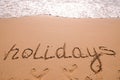 Word Holidays hand written in the sand with a sea wave. Close up sand texture on beach in summer Royalty Free Stock Photo