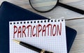 The word PARTICIPATION on a white sheet of paper against the background of a fragment of a notebook and a pen