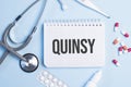 The word quinsy written on a white notepad on a blue background near a stethoscope, syringe, electronic thermometer and pills.