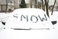 The word `SNOW` is written on the snow-covered windshield of the car. Concept: heavy snowfall in the city, the beginning of winter