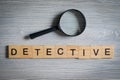 Word of wooden letters - detective, on a gray background with a glass magnifier. Investigation profession concept Royalty Free Stock Photo