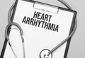 The words Heart arrhythmia is written on white paper on a grey background near a stethoscope. Medical concept