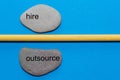 The words hire and outsource are written on natural smooth stones separated by a yellow pencil. The background is isolated in blue