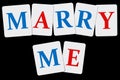 Words marry me composed of letters of blue and red on white cards laid out on a black background