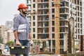 Work day is starting. Cheerful young builder in red helmet holding toolbox, protectives gloves and engineering drawings Royalty Free Stock Photo