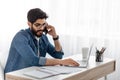 Work from home. Arab freelancer guy talking on cellphone while working with papers and laptop, sitting at home office Royalty Free Stock Photo