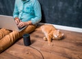 Work from home with funny lazy red cat pet. Man sitting on laminate wooden floor with laptop computer and smart speaker