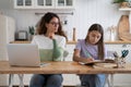 Mother looking at daughter doing homework while working remotely beside, sitting together at table Royalty Free Stock Photo