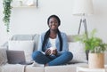 Afro woman freelancer sitting on couch with coffee and laptop