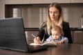 work remotely on maternity leave. Young woman with a baby in her arms happy makes notes in a notebook at a laptop while