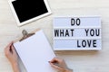 Work space with tablet, female hands, clipboard and `Do what you love` word on modern board over white wooden background, top vi