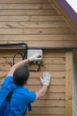 A worker in a blue uniform, connects wires to electricity in the junction box of a wooden house, close-up