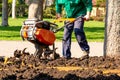 A worker digs the ground in a park around a tree using landscape gardening equipment.