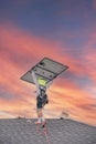 Worker with fall protection rope lifeline, safety harness carrying solar panel walking on shingle roof, wears sun hat neck flap at