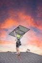 Worker with fall protection rope lifeline, safety harness carrying solar panel walking on shingle roof, wears sun hat neck flap at