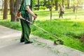 Worker mowing tall grass with electric or petrol lawn trimmer in city park or backyard. Gardening care tools and equipment.