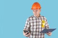 A worker in an orange helmet writes text messages holding a phone and clipboard with papers on a blue background Royalty Free Stock Photo