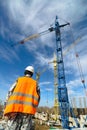 A worker in overalls looks at the high-rise construction cranes on the construction site against a blue sky