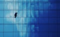 A worker scales the reflective blue glass facade of a modern building, casting a solitary silhouette against the sky.