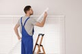 Worker in uniform installing horizontal window blinds on stepladder indoors